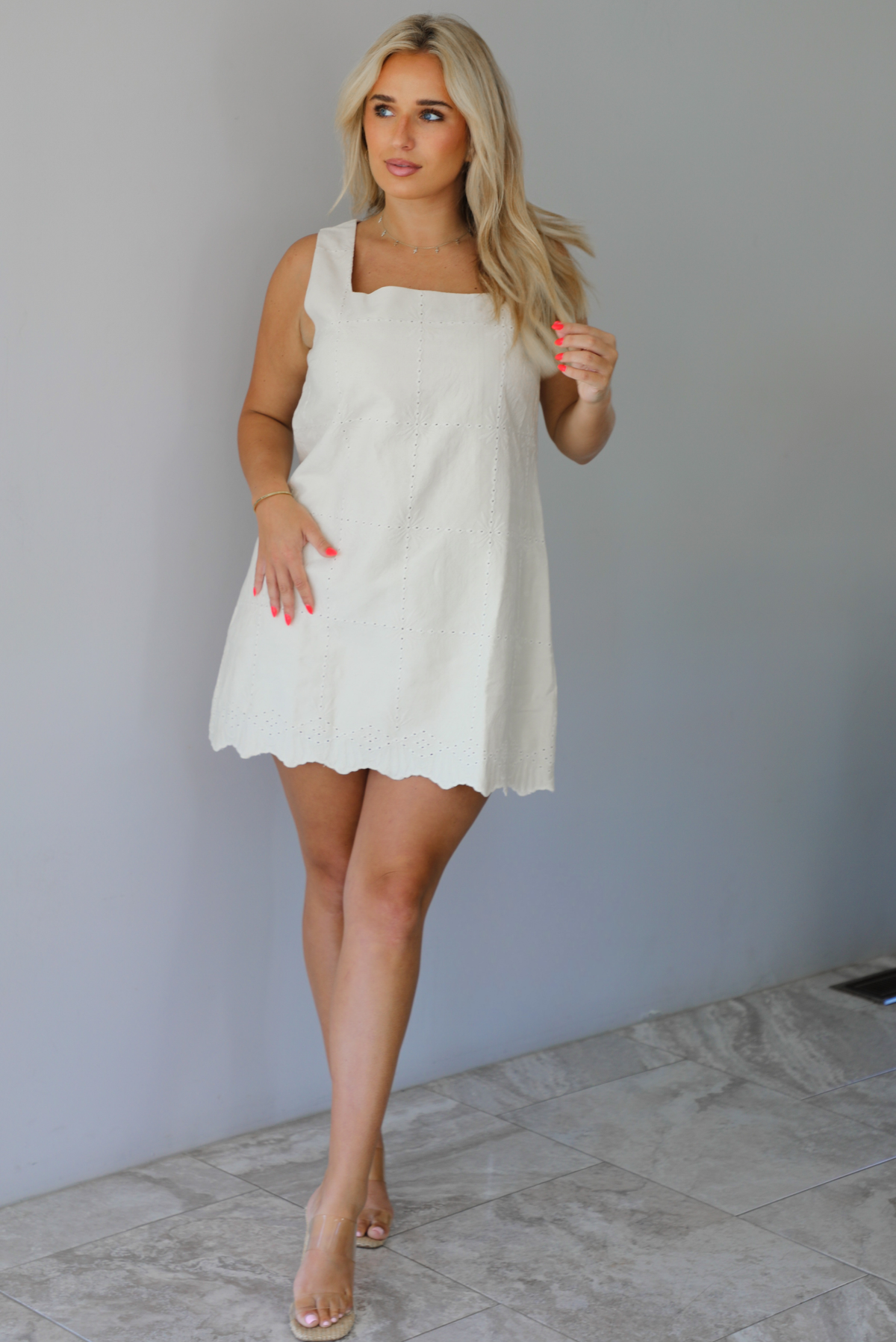 Woman wearing a sand colored mini dress with a square neckline, eyelet details throughout, and a scalloped hem indoors against a white wall with white marble flooring. 