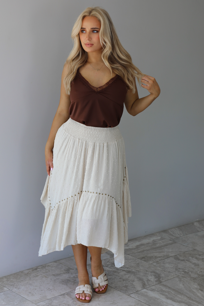 Woman wearing a satin brown tank that is tucked into a high waisted sand colored midi length skirt. The photo is taken in front of a white wall with white marble flooring. 