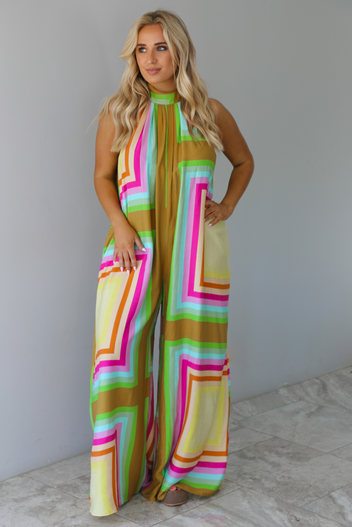 Woman wearing a colorful jumpsuit that has a high neckline, no sleeves, and a flowy cut with flared hem pants. The photo is taken indoors against a white wall with white marble flooring. 