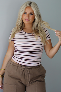 A woman wearing a pink baby tee that has a burgundy and white striped design throughout, paired with mocha barrel denim, standing indoors against a white wall.