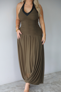 A woman wearing an olive maxi dress with black trim detailing at the v-halter neckline, has a fitted bodice and drop waist design, with a maxi-length flowy skirt, standing indoors against a white wall with white marble flooring. The photo shows the dress without showing the models face. 