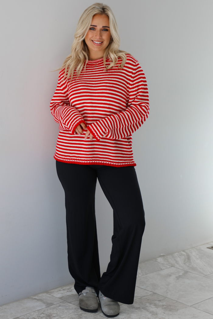 Woman wearing a white and red striped sweater that has a high neckline, an oversized fit, and is paired with black leggings. The photo is taken indoors against a white wall with white marble flooring. 