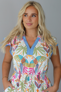 A woman wearing a short sleeved midi dress with a colorful tropical leaf print and light blue trim indoors against a white wall. The photo shows a closeup of the bodice of the dress, showing the v-neckline. 