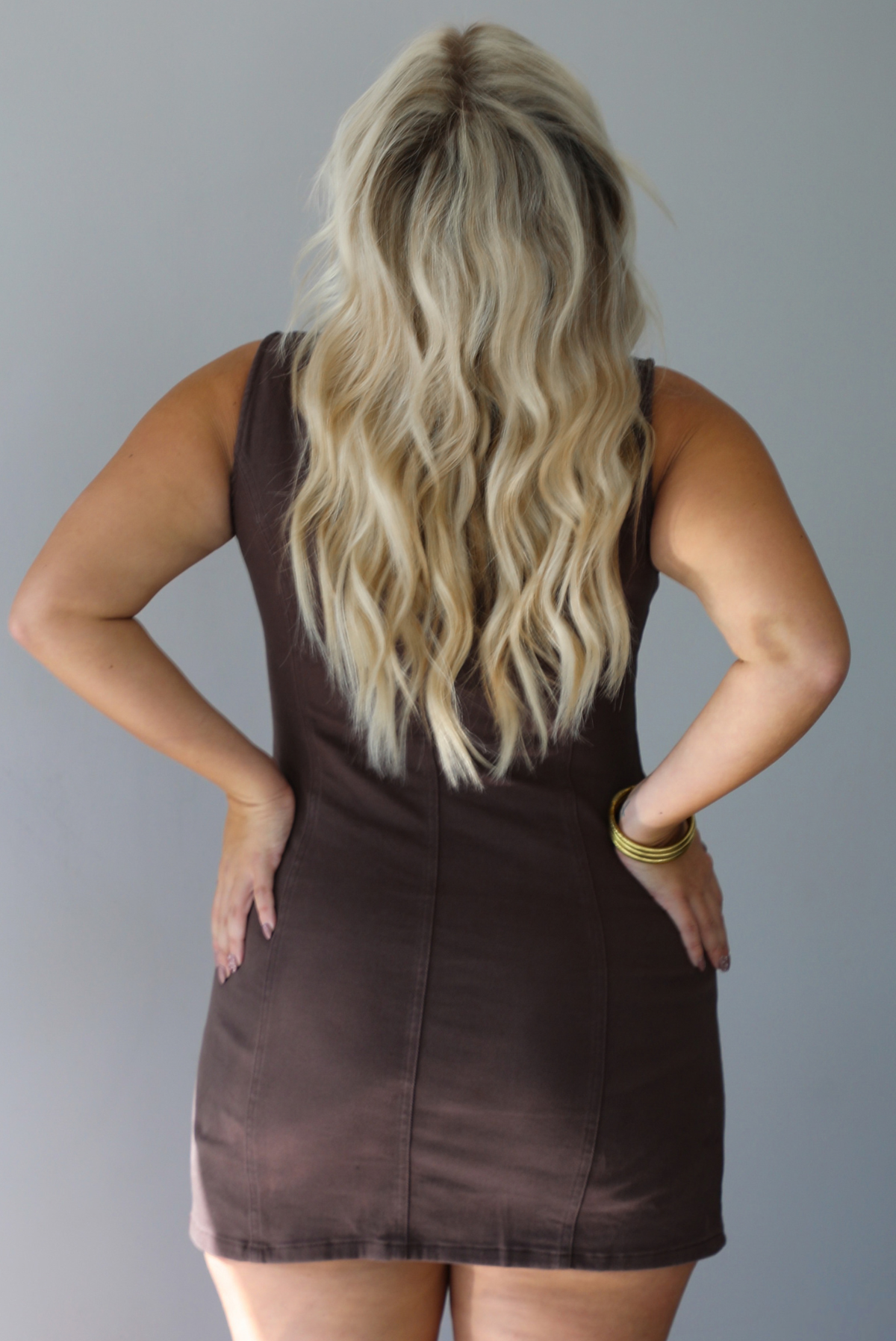 A woman shown from behind modeling a sleeveless brown mini dress with a button-front closure and tie neckline, standing indoors against a white wall.