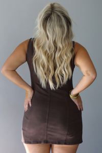 A woman shown from behind modeling a sleeveless brown mini dress with a button-front closure and tie neckline, standing indoors against a white wall.
