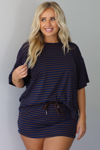 A woman wearing a navy and brown striped set featuring an oversized top and mini skirt, standing indoors against a white wall.