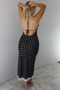 Woman wearing a black wearing a white polka dot printed midi dress that has a halter tie neckline, is form fitting, with white lace trim at the bottom. The photo is taken indoors against a white wall and shows the back of the dress, which is open and ties in the center of the back.