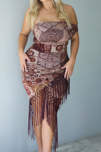 A woman wearing a burgundy and multi-colored patterned dress with a faux-wrap design, off the shoulder one sided sleeve, and fringe detailing at the hem, standing indoors against a white wall.