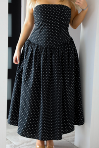 Close up of a woman model wearing a strapless/sleeveless, black with white polka dots, maxi length dress indoors. It has a fitted bodice with a flowy skirt. The photo showcases the dress in its entirety.