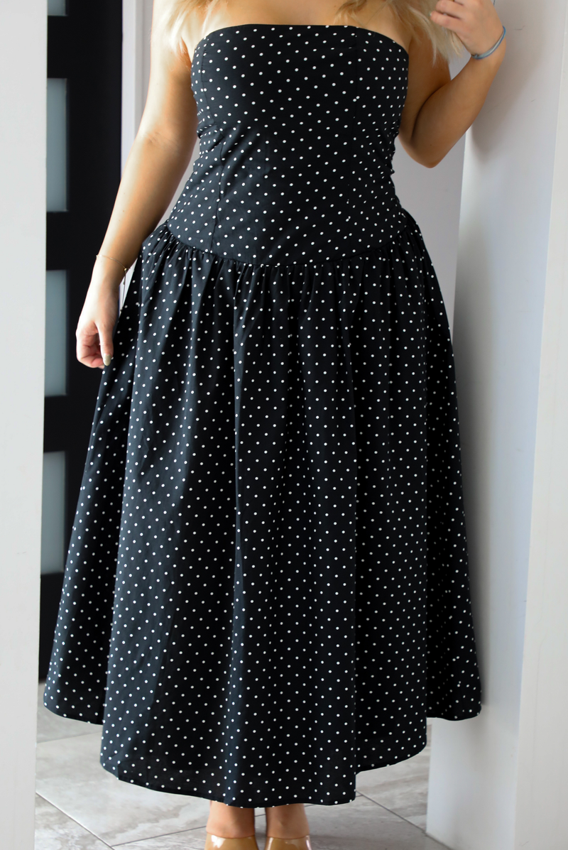 Close up of a woman model wearing a strapless/sleeveless, black with white polka dots, maxi length dress indoors. It has a fitted bodice with a flowy skirt. The photo showcases the dress in its entirety.