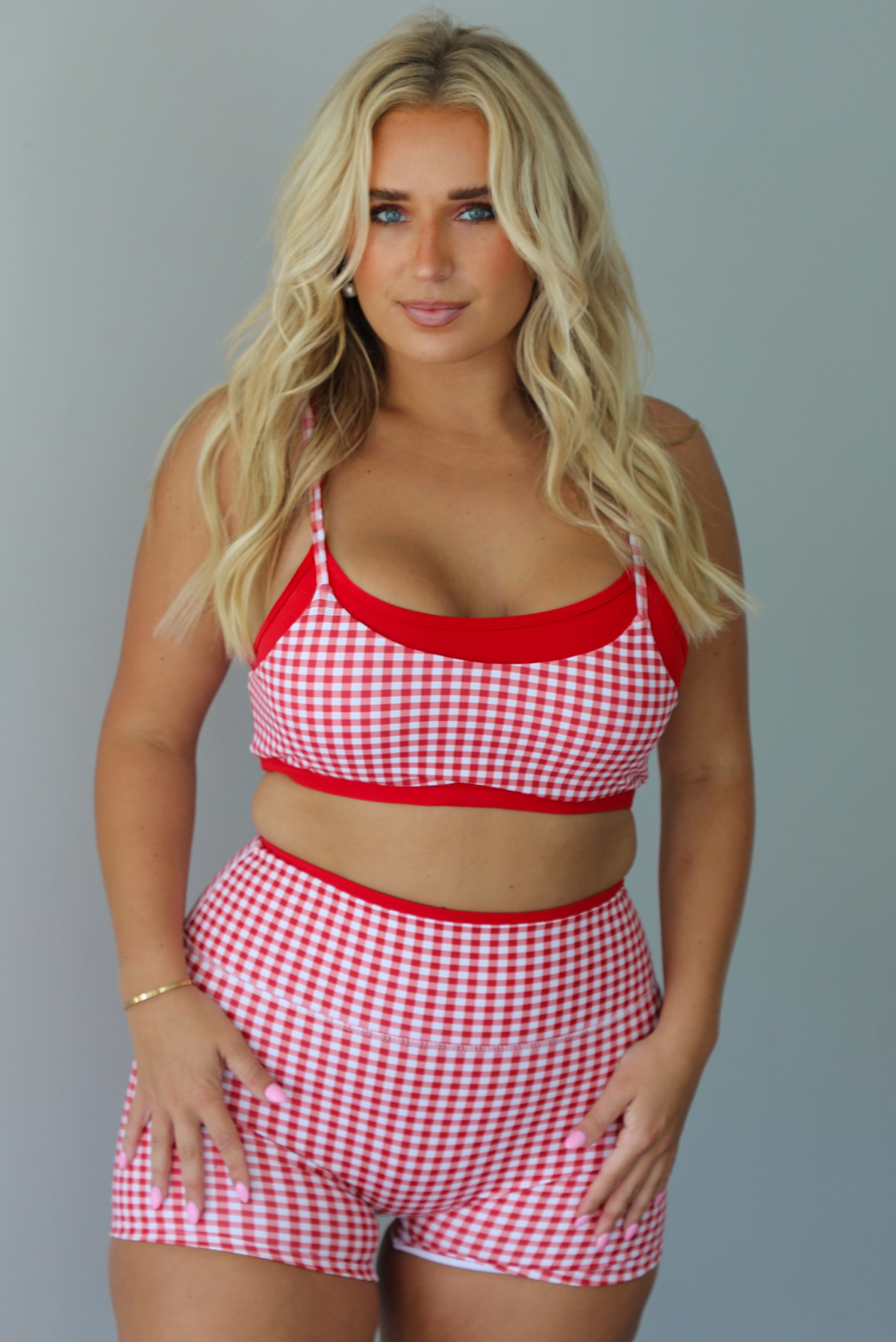 A woman modeling a red and white gingham two-piece athletic set featuring a sports bra and high waisted biker shorts with solid red trim indoors against a white wall.