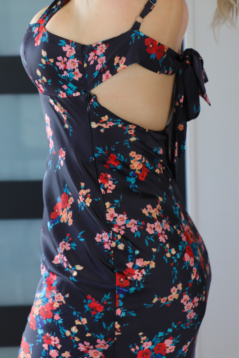 Close up of the side of a woman model wearing a navy dress with a multi-colored floral print. The dress is a satin material, is a midi length, and is curve-hugging. The photo is taken inside a modern white and black interior. The photo shows the zipper closure on the side. 