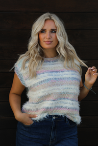 Woman model wearing a soft, striped sweater vest with dark wash denim jeans. The photo showcases a closeup of the sweater vest. The photo is taken outdoors against a dark brown wooden door. 