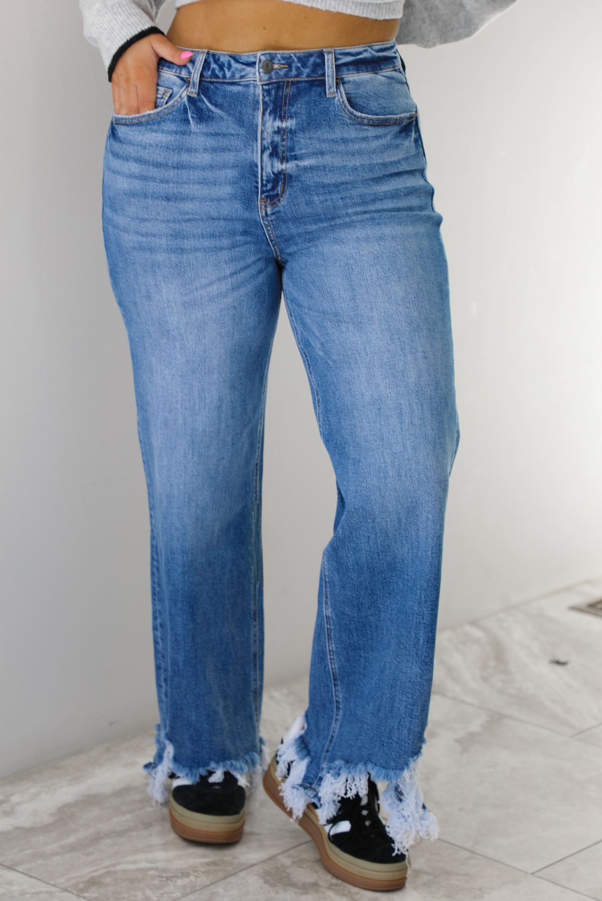 Woman wearing a medium wash denim that are high waisted, have functional front and back pockets, functional button and zipper closures, and have a raw, distressed hem. The photo is taken indoors against a white wall with white marble flooring.