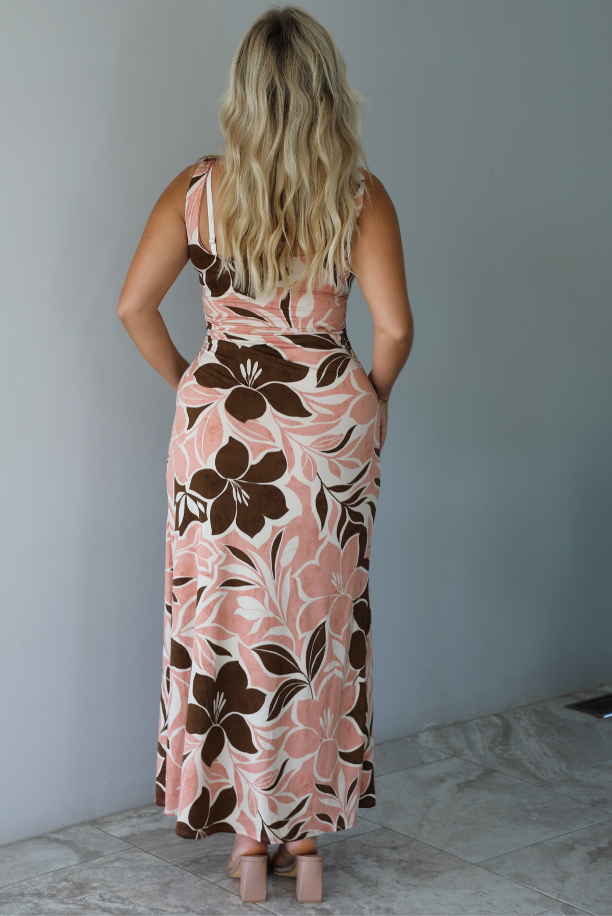A woman modeling a thick strapped maxi dress with a pink, brown, and white floral pattern, standing indoors against a white wall with white marble flooring. The photo shows the back of the dress. 