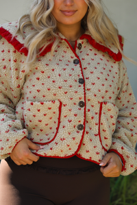 Woman model wearing a tan and red sherpa cardigan with a collared neckline and red trim with dark brown ahtleisure pants. The photo is taken outdoors with a white wall and greenery in the back. The photo showcases a closeup of the sherpa jacket with the functional buttons and pockets in the front. 