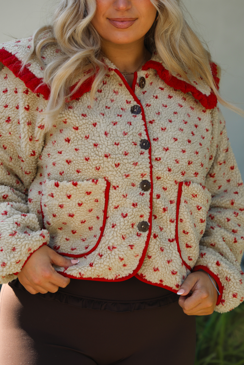 Woman model wearing a tan and red sherpa cardigan with a collared neckline and red trim with dark brown ahtleisure pants. The photo is taken outdoors with a white wall and greenery in the back. The photo showcases a closeup of the sherpa jacket with the functional buttons and pockets in the front. 