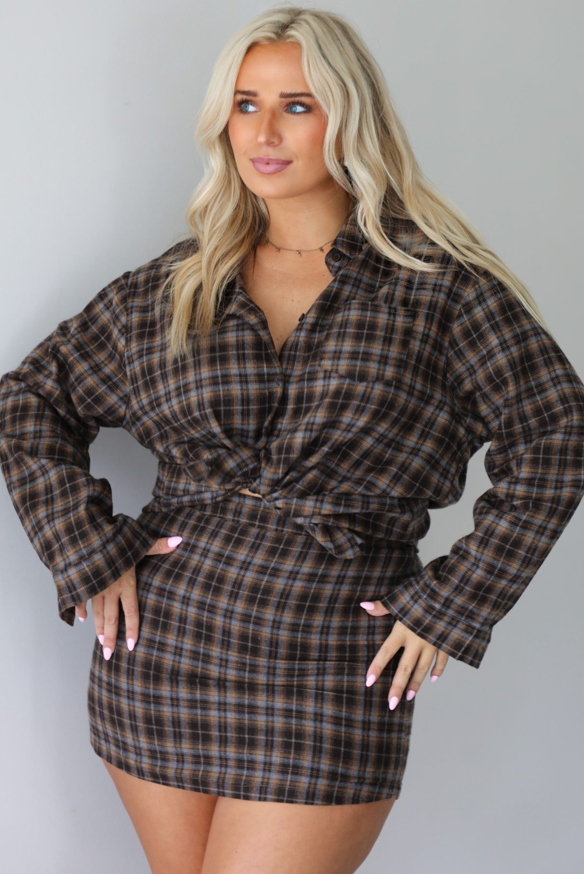 A woman wearing aa dark brown plaid set featuring an oversized button-down and fitted mini length skirt, shown with the top tied in the front giving it a cropped look, standing indoors against a white wall.