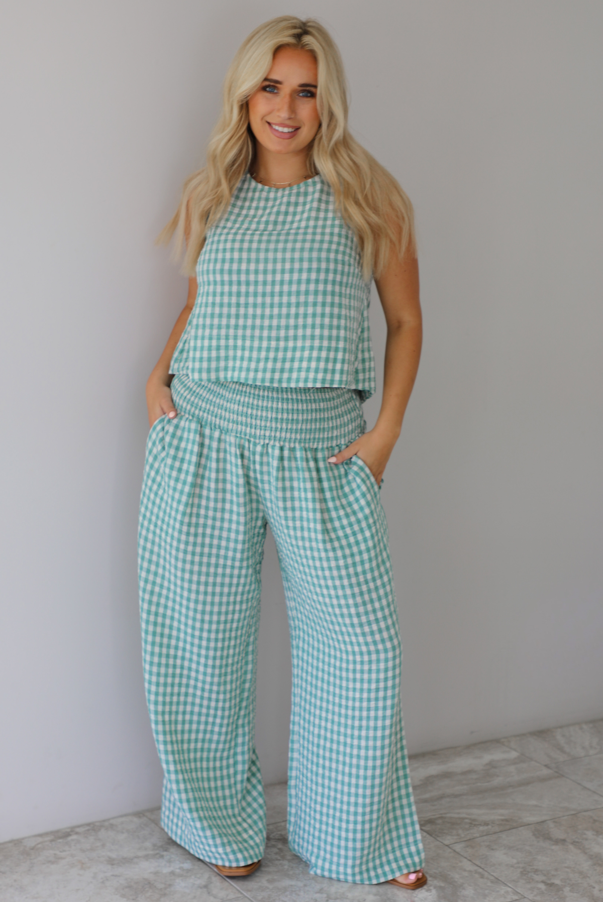 Woman wearing a mint and white gingham set with an oversized top and flared hem pants indoors against a white wall with white marble flooring. 