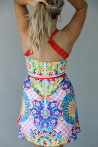 Woman wearing a multi-colored and multi-patterned mini dress with thick red straps, a cinched waistline with a removable tie, and a flowy skirt indoors against a white wall. The photo shows the back of the dress, which has crisscross straps, a smocked back, and a zipper closure in the back. 