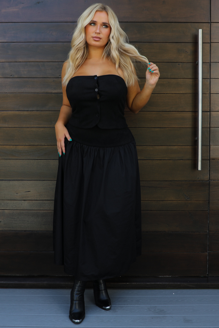 Woman model wearing a black two piece set that features a strapless tube style top that has functional button closures in the front, and a high waisted maxi length skirt. The set has contrasting textures, with the top being a suede texture, and the skirt having the same suede texture at the waistline with a stiffer cotton feel below the hips. The set is paired with black, silver tipped booties. The photo is taken outdoors against a dark brown wooden door.