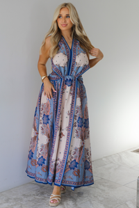 Woman wearing a blue and multi-colored halter tie maxi dress that has a long, flowy skirt with a split hem detail. The photo is taken indoors against a white wall with white marble flooring. 
