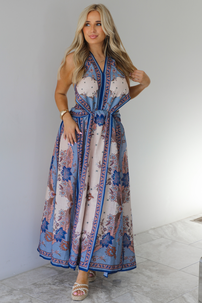 Woman wearing a blue and multi-colored halter tie maxi dress that has a long, flowy skirt with a split hem detail. The photo is taken indoors against a white wall with white marble flooring. 