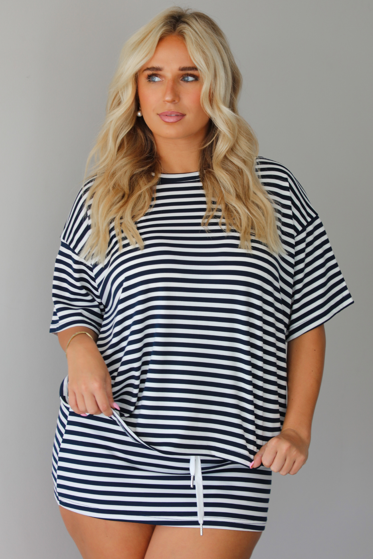 A woman wearing a navy and white horizontally striped short-sleeve oversized tee and skirt with a white drawstring tie at the waist, standing indoors against a white wall.