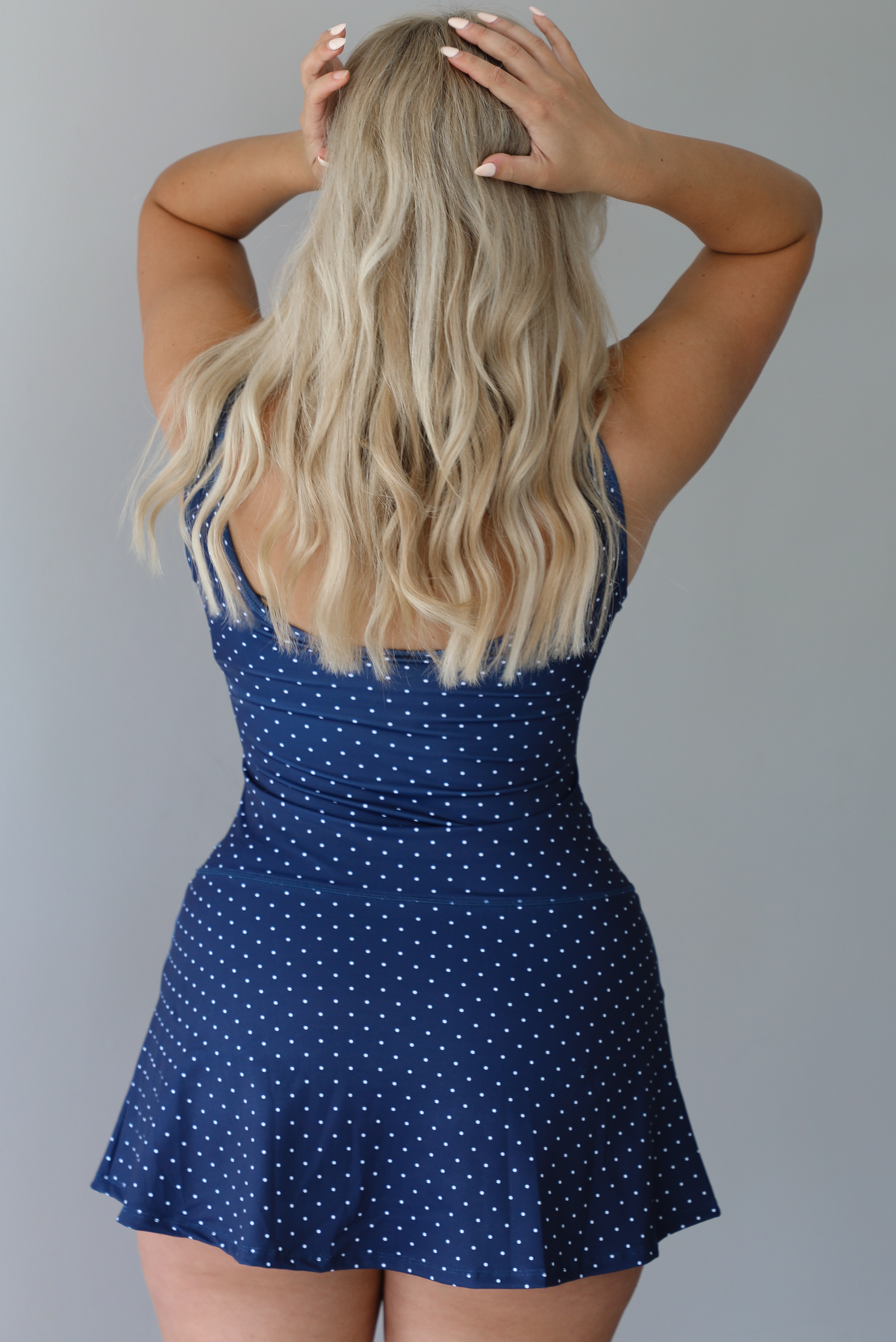 Woman wearing a navy and white polka for athleisure dress indoors against a white wall. The photo shows the back of the dress. 
