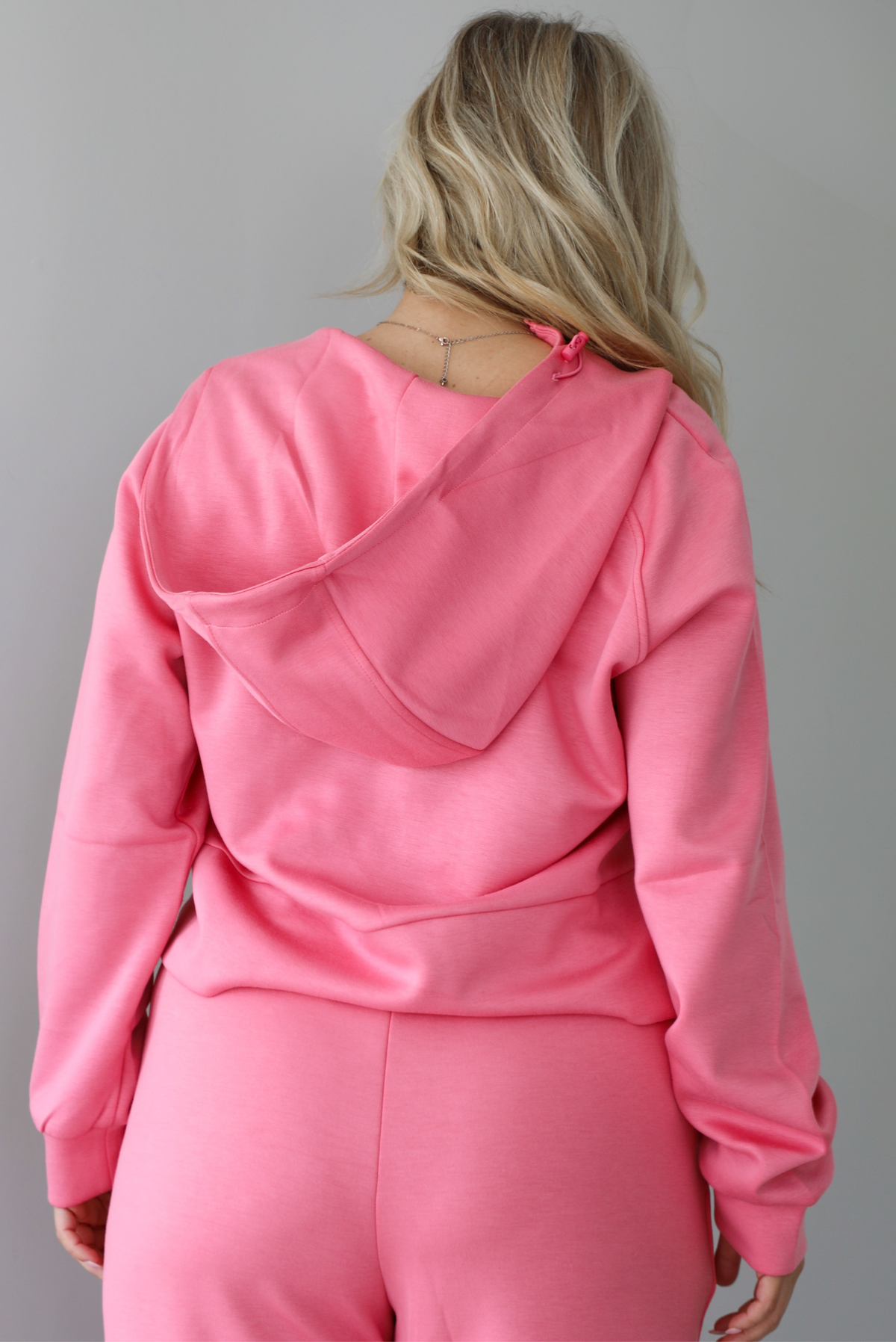 Woman wearing a pink sweat-set featuring a cropped zip-up hoodie with pants. The top of the set has a hoodie detail, a functional zipper closure in the front, and a cropped cut with an open detail under the zipper. The photo shows the back of the top. The photo is taken indoors against a white wall.