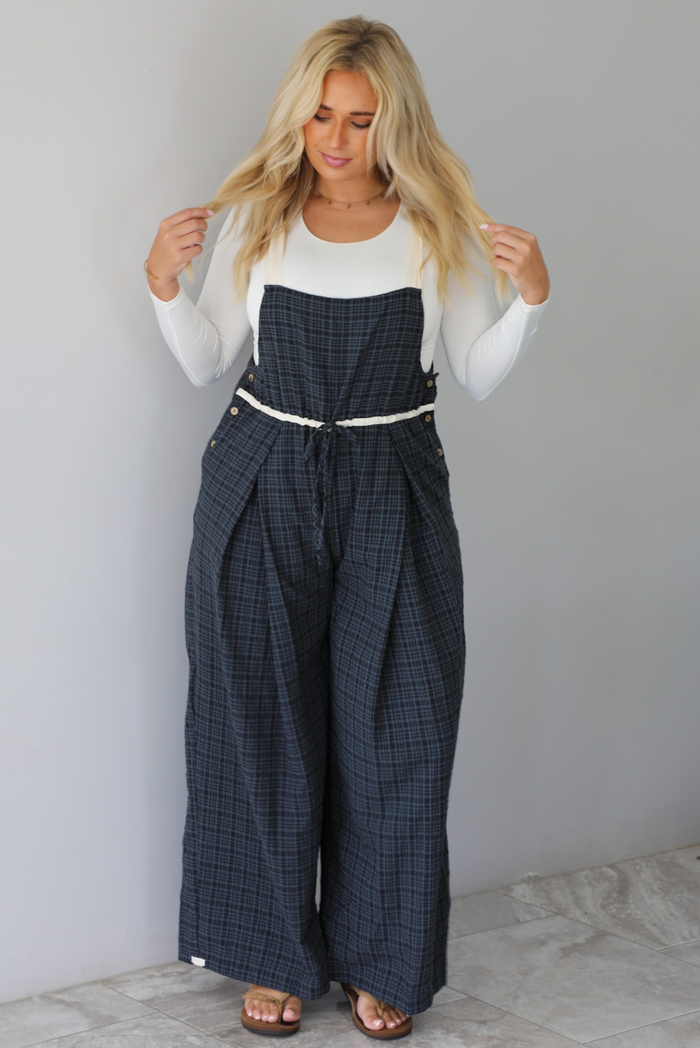 A woman modeling a white long-sleeve top and dark gray plaid wide-leg overalls, standing indoors against a white wall with white marble flooring. 