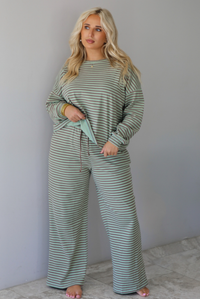 A woman wearing a pale green and brown striped two piece set featuring an oversized long sleeved top and high waisted, wide leg pants, standing indoors against a white wall with white marble flooring. 