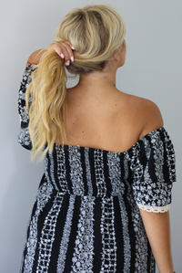 A woman shown from behind wearing a black and white maxi dress with a white floral print that alternates between a cluster appearance and a striped appearance, with off the shoulder sleeves and a flowy skirt, standing indoors against a white wall. The photo shows a closeup of the back of the dress with the smocked detail. 