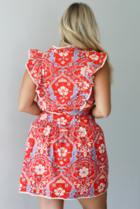 A woman modeling a sleeveless mini dress with a red, cream, and light blue paisley pattern, featuring a tie waist and ruffled collar, standing indoors against a white wall. The photo shows the back of the dress. 
