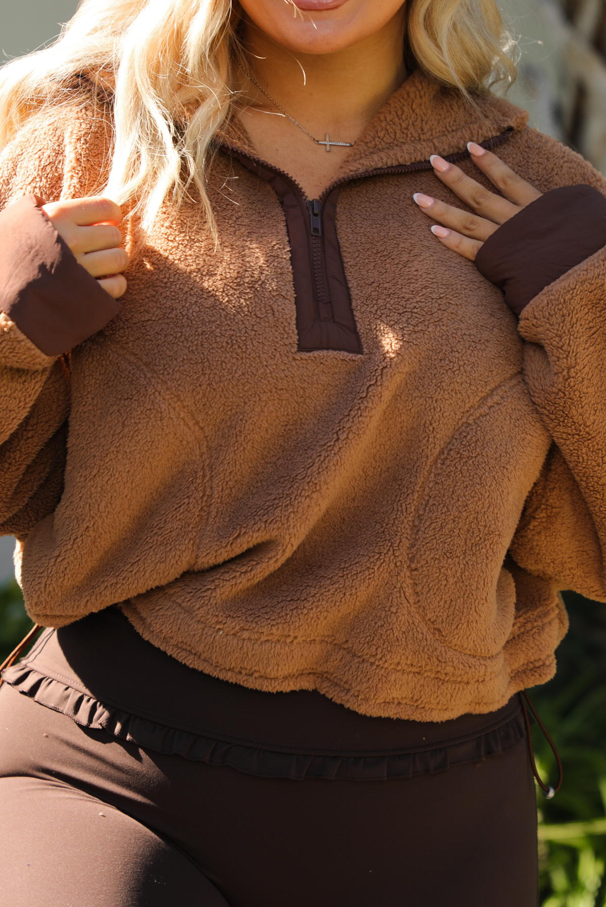 Woman model wearing a tan and brown quarter zip sweater with dark brown athleisure pants. The photo is taken outdoors against some greenery and a white wall. The photo showcases the zipper closure in the front. 