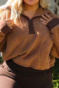 Woman model wearing a tan and brown quarter zip sweater with dark brown athleisure pants. The photo is taken outdoors against some greenery and a white wall. The photo showcases the zipper closure in the front. 