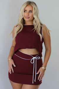 A woman in a two-piece burgundy set consisting of a sleeveless top that's bra tucked giving it a cropped look and a matching mini skirt with white trim detailing, and a tie waist detail, paired with black pointed-toe heels, standing indoors against a white wall.
