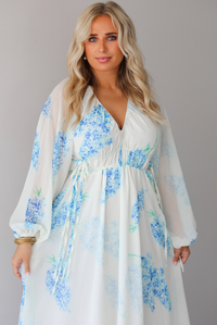 Closeup of a woman modeling a white maxi dress with a blue floral print and a sheer, flowing overlay, focusing on the bodice of the dress with its cinched waistline, standing indoors against a white wall. 