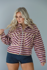 Woman wearing a pink, burgundy, lilac, and sand colored jacket with a charcoal colored two-piece set featuring a cropped sports bra and high waisted short biker shorts. The photo shows the jacket zipped up in the front with the functional pockets in the front. The photo is taken indoors against a white wall.