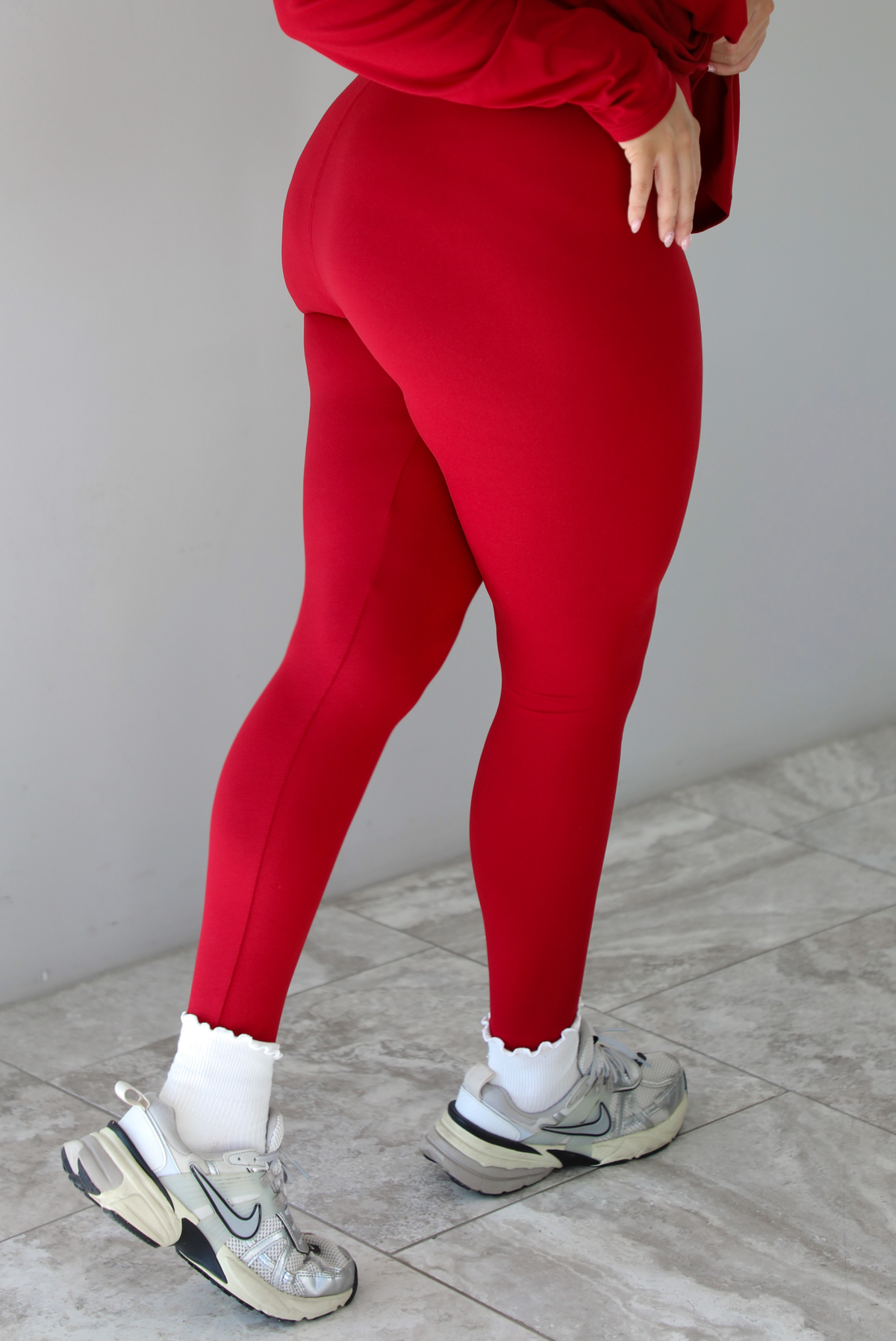 Woman wearing a red two piece set that features an oversized long sleeved top and fitted leggings. The photo shows a closeup of the back of the pants that are paired with high white ankle socks and silver sneakers. The photo is taken indoors against a white wall with white marble flooring.