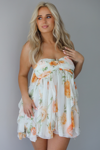 Woman wearing a white, strapless, babydoll cut dress that has an orange and green floral print pattern and flowy skirt indoors against a white wall.