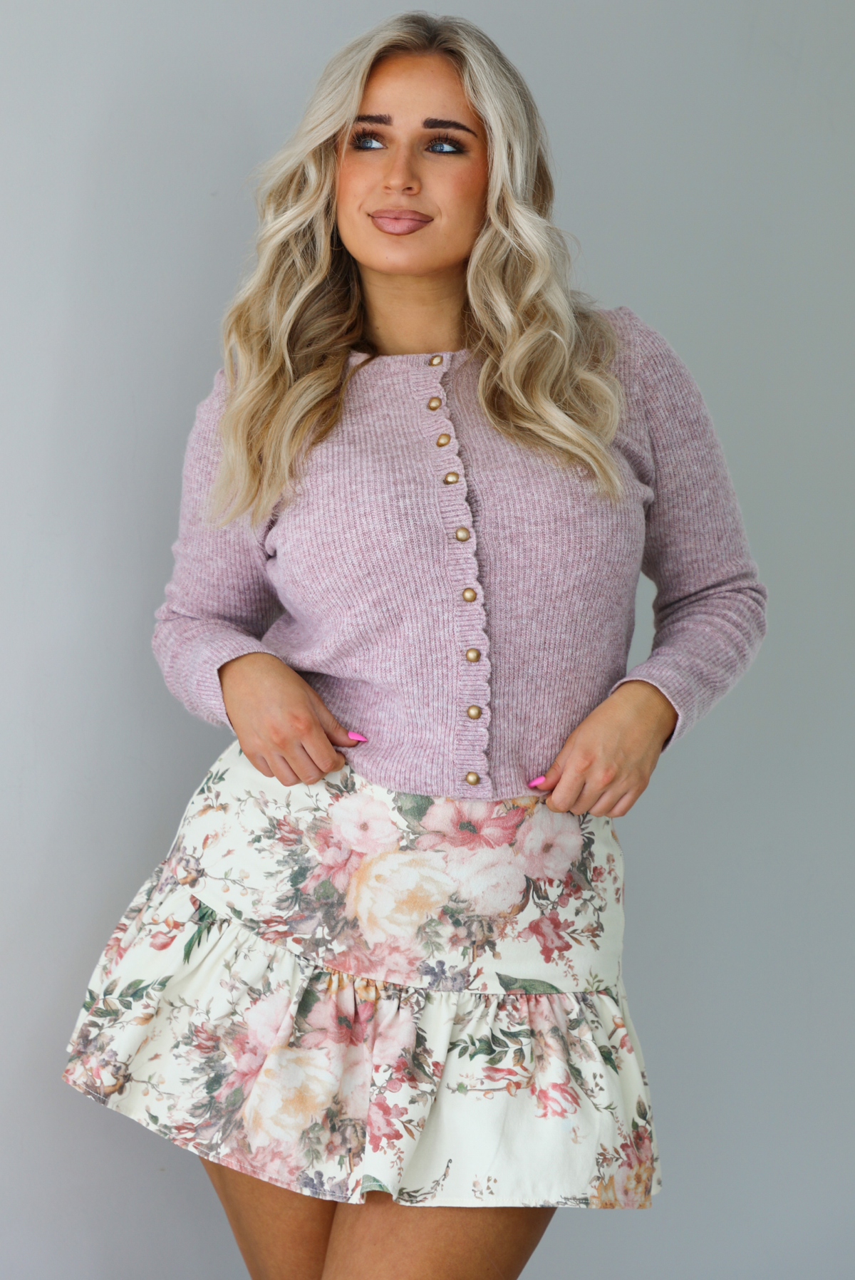 Woman wearing a mauve colored ribbed cardigan that has gold button closures along the front with a cream and multi-colored floral print patterned skirt. The photo is taken indoors against a white wall.