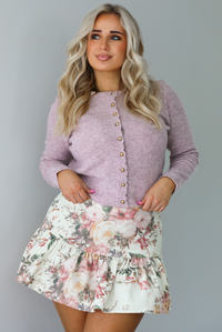 Woman wearing a mauve colored ribbed cardigan that has gold button closures along the front with a cream and multi-colored floral print patterned skirt. The photo is taken indoors against a white wall.