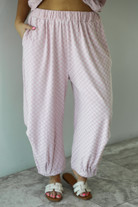 Woman wearing a pink checkered set featuring a slouchy top and pants. The photo is taken indoors against a white wall and shows a closeup of the pants, which are high waisted, have an elastic waistline, functional pockets on either side, have an oversized fit, and cinch slightly with elastic at the hem of the pants.