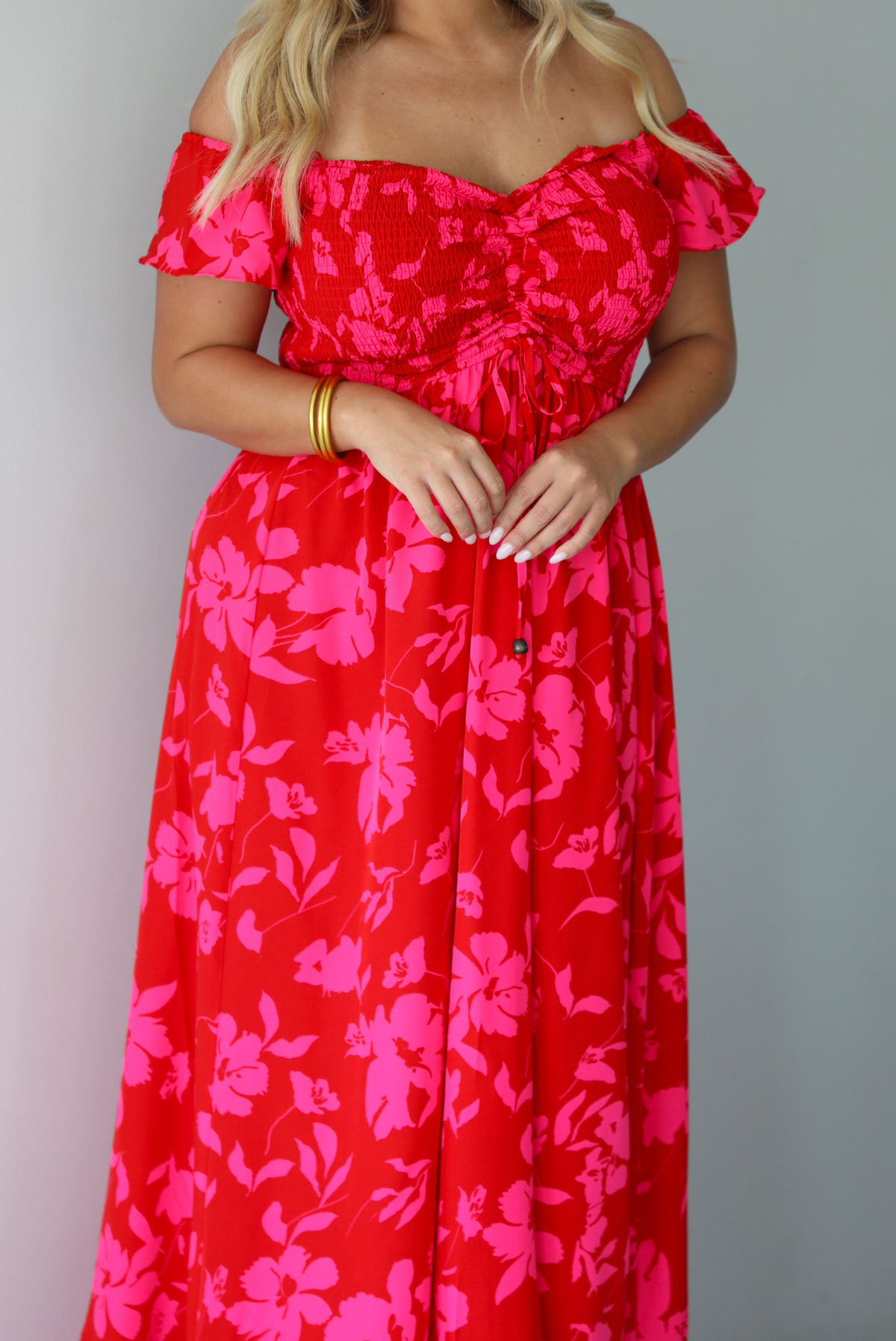 A closeup of a woman modeling a bright red off-the-shoulder maxi dress with a vibrant pink floral pattern, focusing in on the smocked bodice and drawstring detail in the center of the bodice, standing indoors agains a white wall.