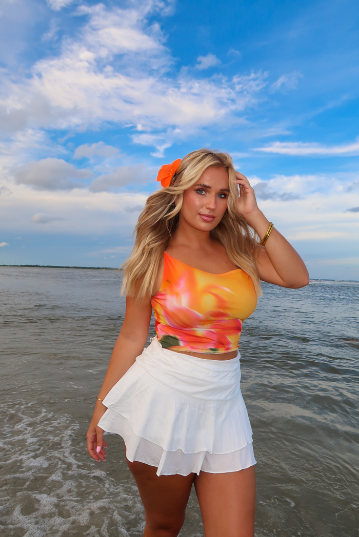 A woman stands on a beach wearing a bright tie-dye crop top in orange, yellow, and white, paired with a white ruffled skirt. She has a bright orange flower in her hair and is posing under a blue sky with clouds.