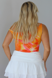 A woman wearing a bright tie-dye crop top in orange, yellow, and white, paired with a white ruffled skirt indoors against a white wall.  The photo shows the back of the top. 