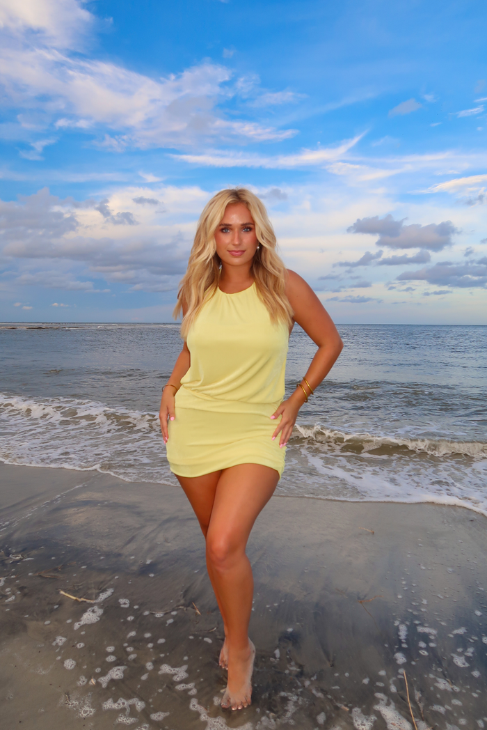A woman stands on a beach wearing a yellow sleeveless mini dress, with ocean waves and a blue sky in the background.
