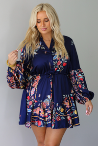 A woman modeling a navy blue mini dress with a colorful floral print and long balloon sleeves, paired with beige ankle boots, standing indoors against a white wall.