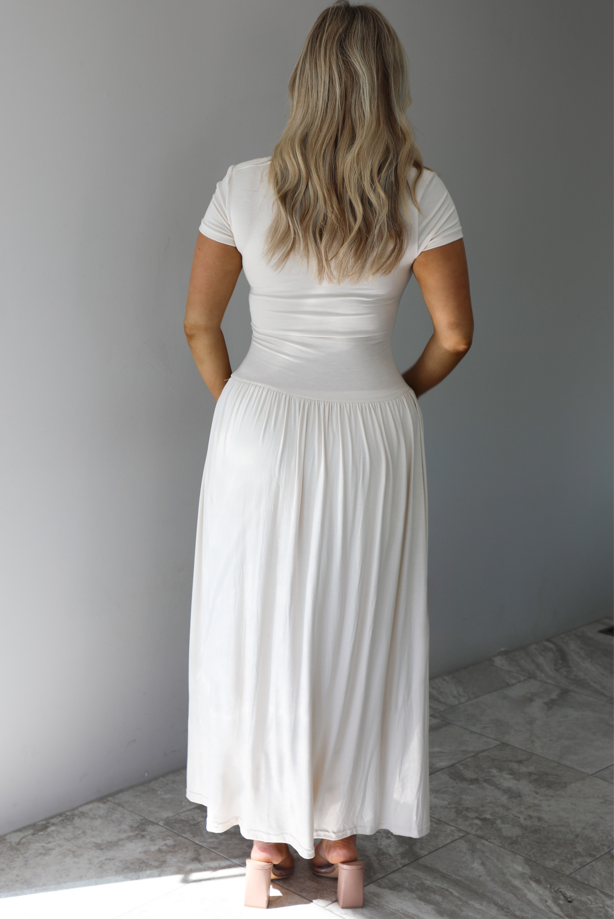 Woman wearing a light sand colored maxi length dress that has a fitted bodice with lace detailing in the center of the bust, a drop waist design, short sleeves, and a flowy skirt. The photo is taken indoors against a white wall with white marble flooring. The photo shows the back of the dress. 